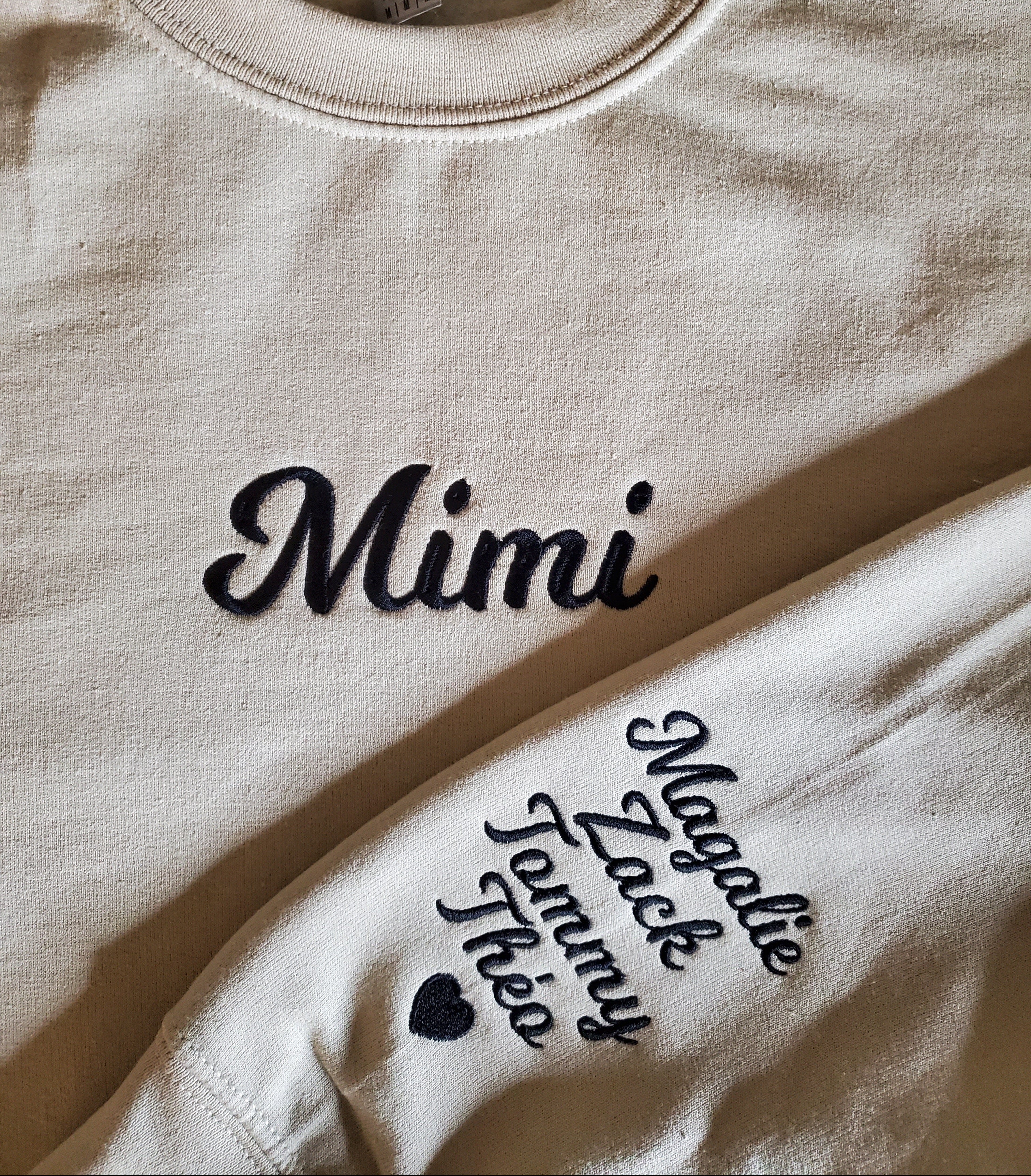 Beige sweatshirt with 'Mimi' and additional text in black on a plain background