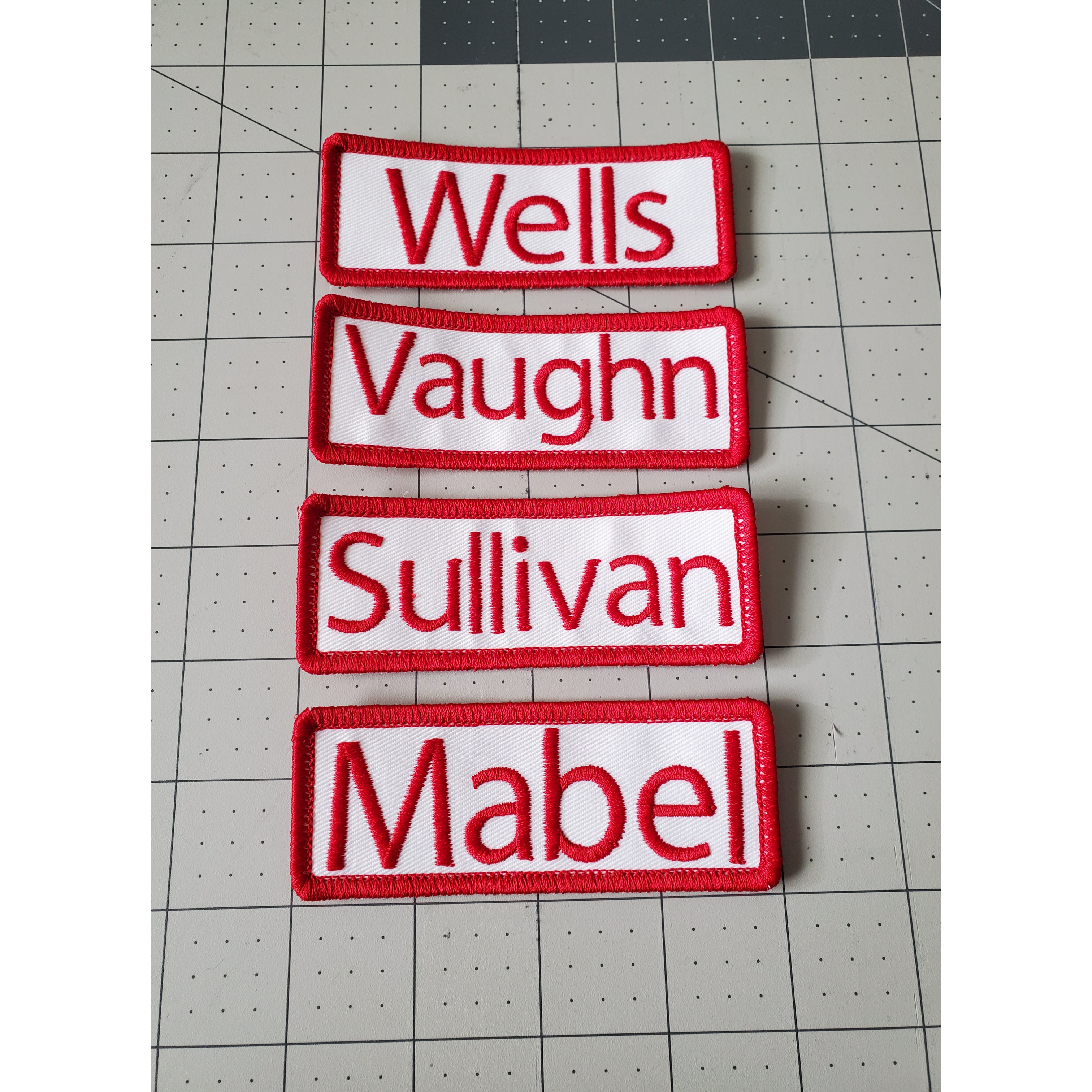 Four rectangular name patches with red text on a white background, placed on a grid surface.